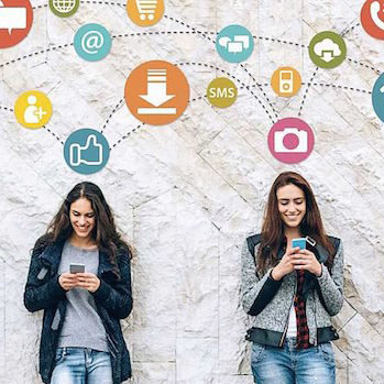 two girls leaning against a wall and looking at their smartphones